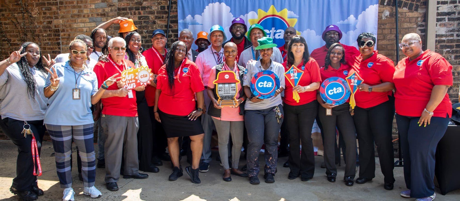 CTA employees posing for a group picture. Some are wearing red shirts with the CTA logo or bucket hats. A few hold signs with the logo, a train, or "fun in the sun." 