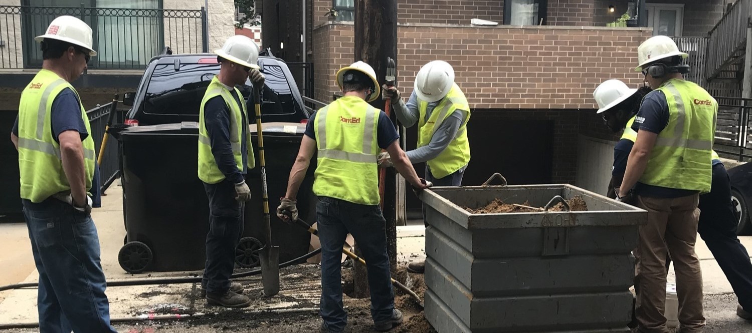 A construction crew in yellow work vests performing utility work around the base of an electrical pole.