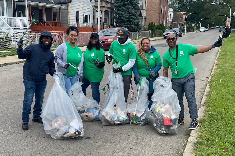 CTA employees in green shirts smiling and holding trash bags while they clean up a neighborhood.